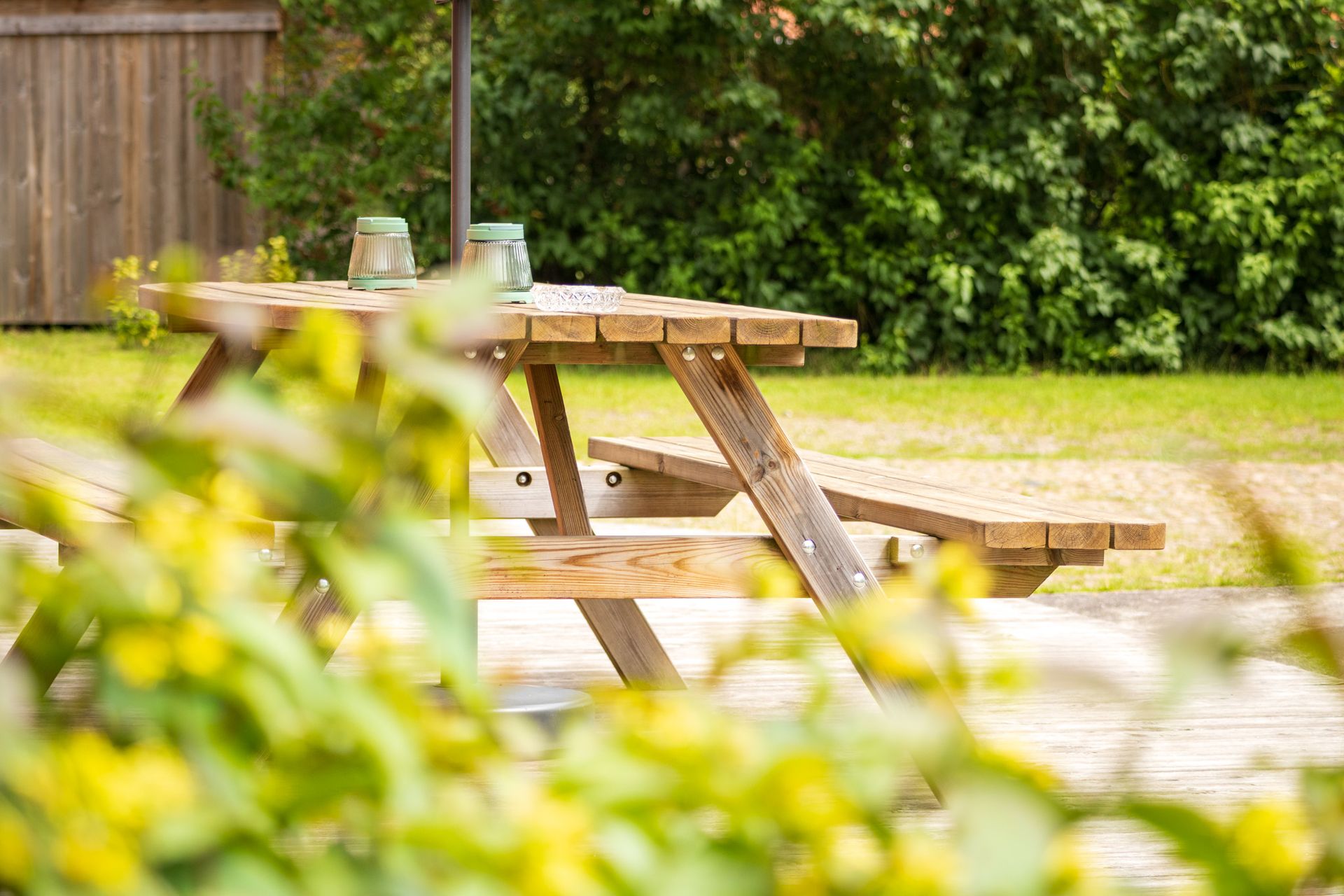 Ein hölzerner Picknicktisch in einem sonnigen Garten, im Hintergrund Grün und ein Zaun.