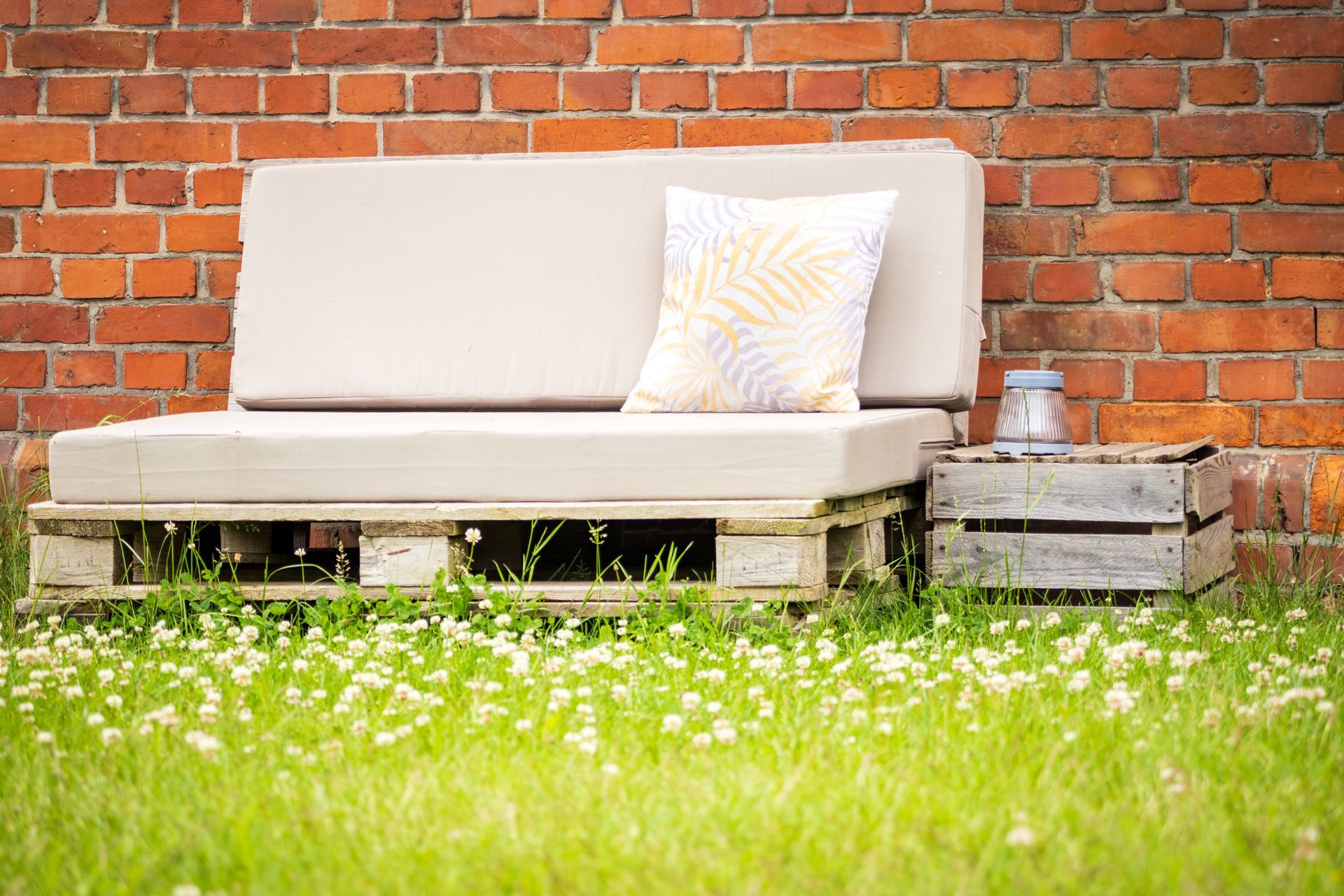 Graues Sofa im Freien mit gemustertem Kissen auf Paletten vor einer Backsteinmauer, daneben ein Kistentisch im Gras