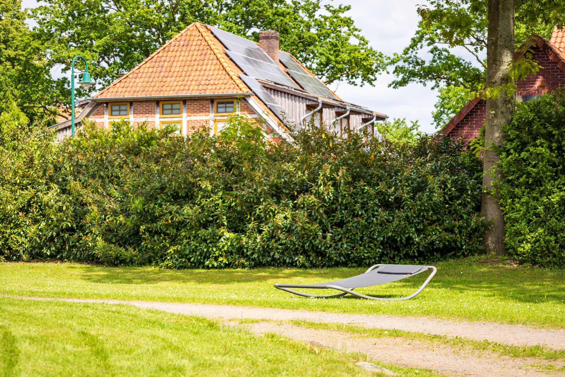 Das Haus ist teilweise hinter einer Hecke versteckt, auf einem sonnigen Rasen steht ein Liegestuhl.