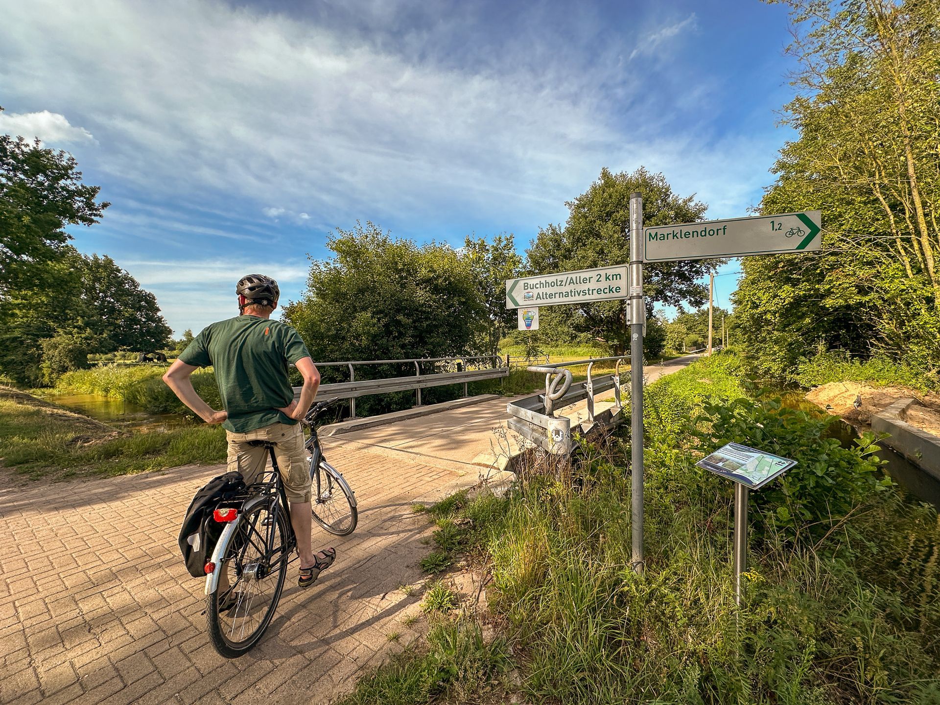 Ein Radfahrer mit Helm und grünem Hemd steht auf einem gepflasterten Weg neben einem Wegweiser in sonniger Umgebung.