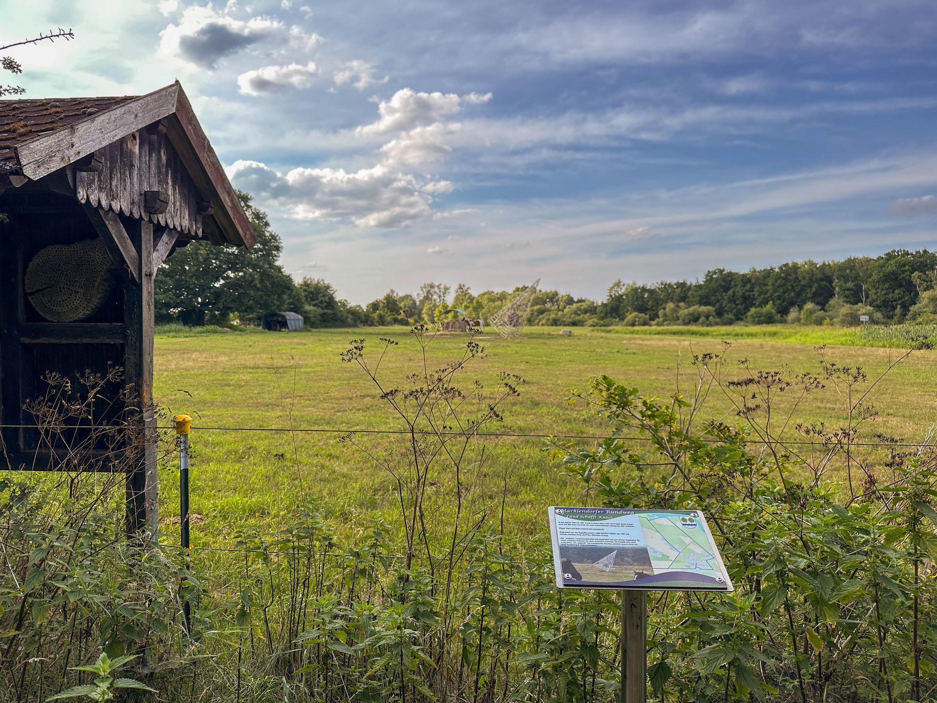 Eine grasbewachsene Wiese mit einem hölzernen Informationskiosk und einem kleinen Schild vor dem Hintergrund von Bäumen.