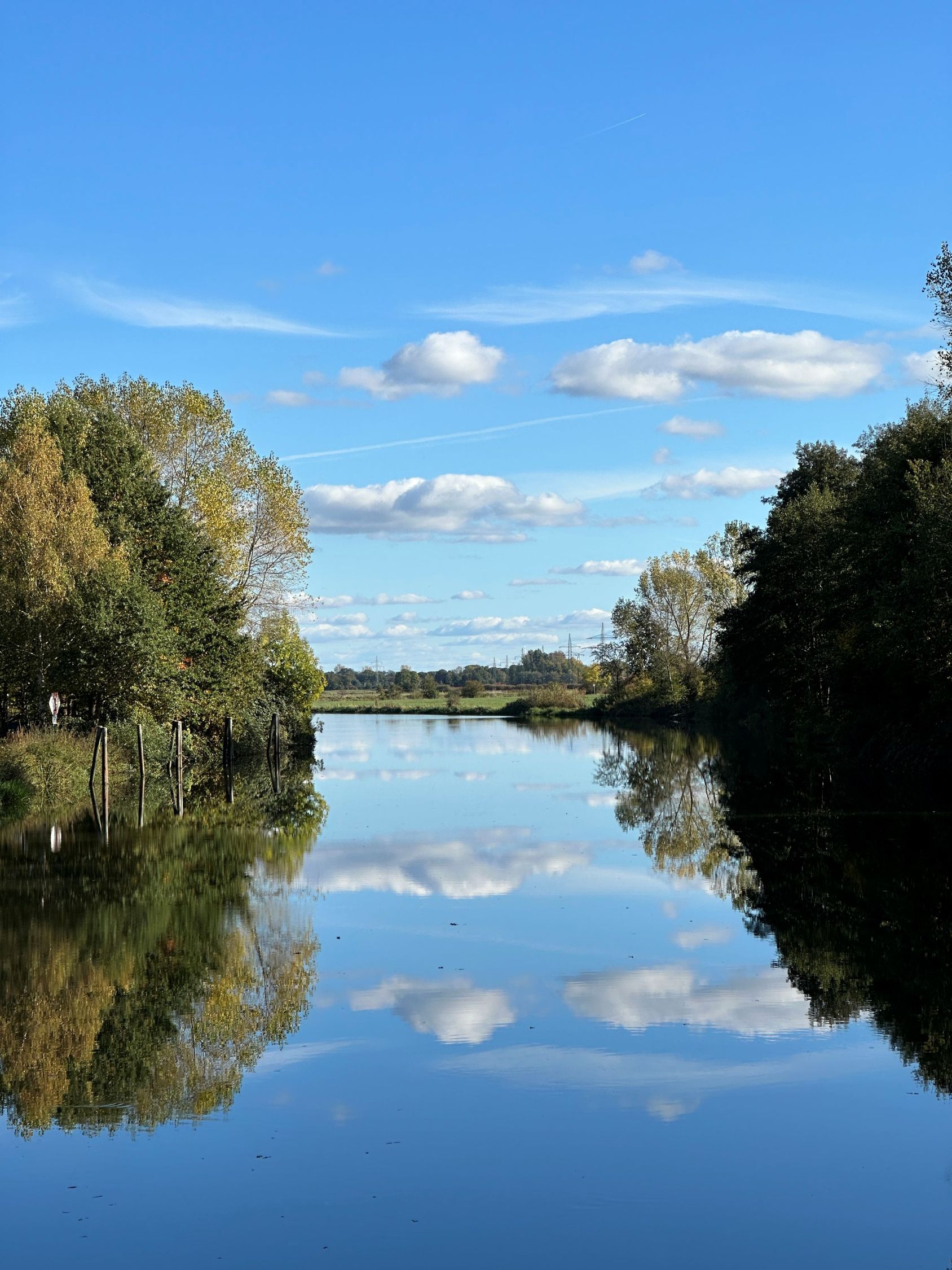 Ein ruhiger Fluss, in dem sich ein blauer Himmel und weiße Wolken spiegeln, eingerahmt von grünen Bäumen an den Ufern.