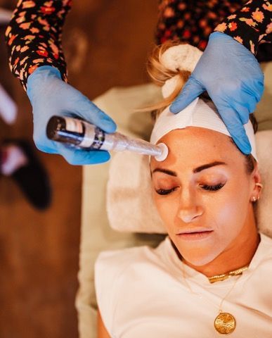 Person receiving a facial treatment; device held above forehead. Blue gloves, white cloth, gold jewelry.