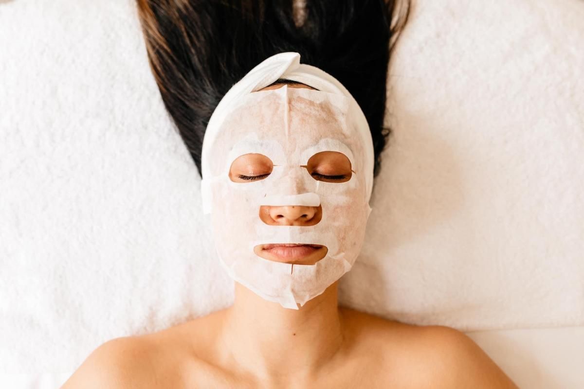 Person wearing a white sheet mask with eye, nose, and mouth cutouts, lying on white surface.