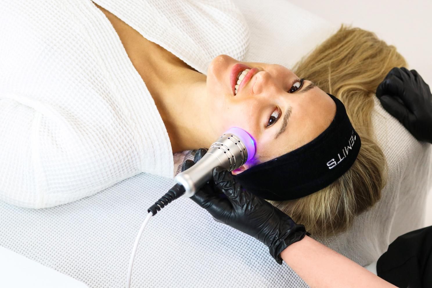 Woman receiving facial treatment with a handheld device, under bright light. She smiles while lying down in a spa.
