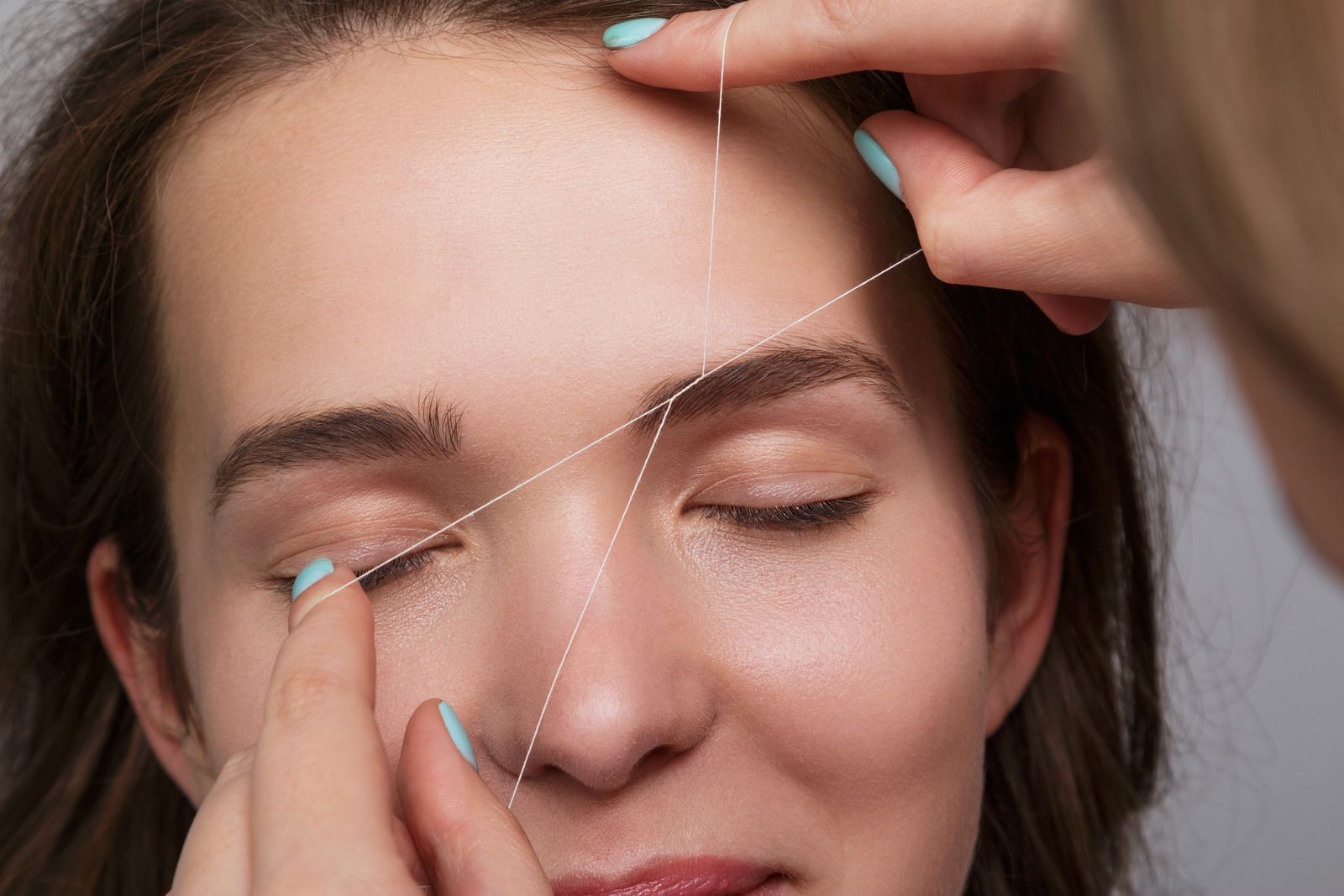 Person's face with closed eyes, getting eyebrows threaded with white string. Hands are in frame.