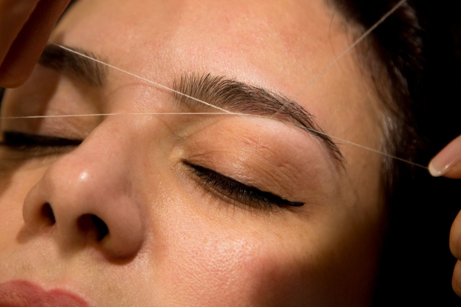 Eyebrow threading: a person's eye closed, thread held near eyebrow, two hands, close-up.