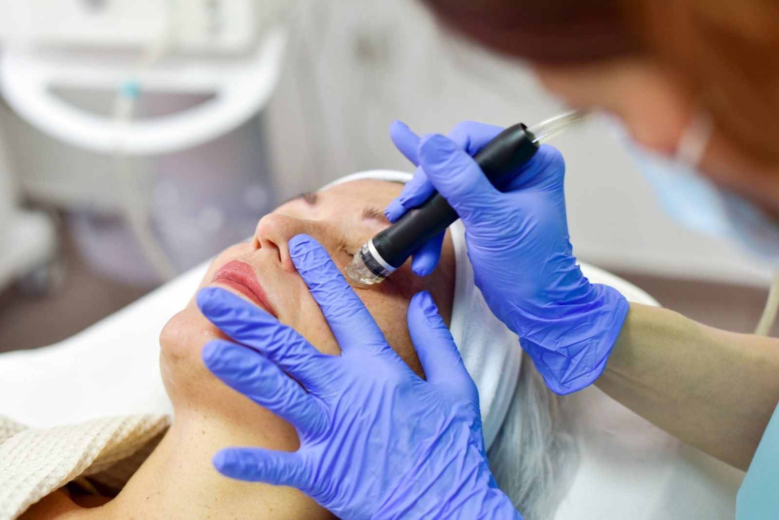 Woman receiving facial treatment; cosmetologist uses a device near her eye area. Blue gloves, white towel, clinical setting.