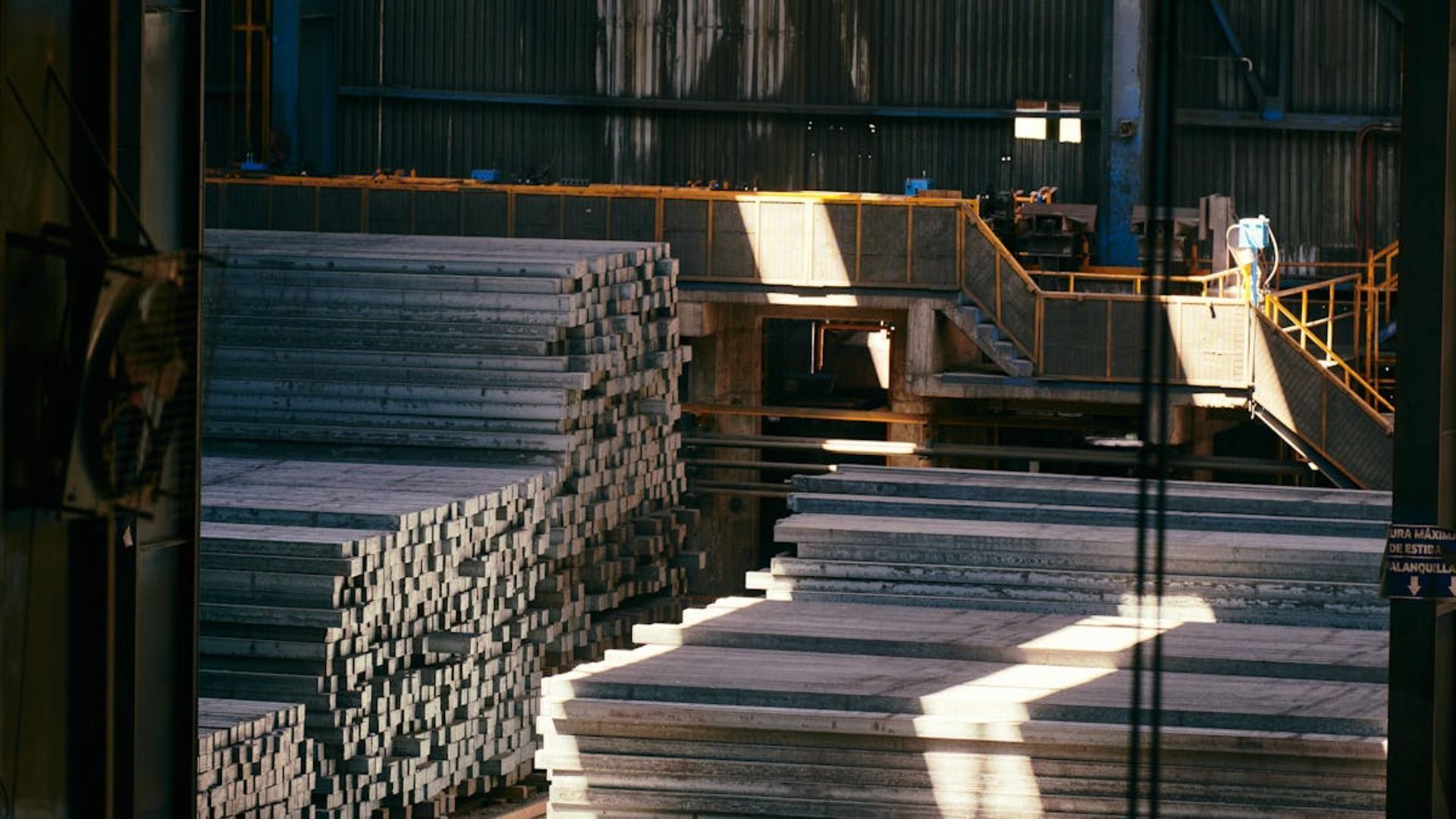 Barras de metal apiladas y losas industriales rectangulares llenan un espacio amplio y con poca luz, propio de un almacén.