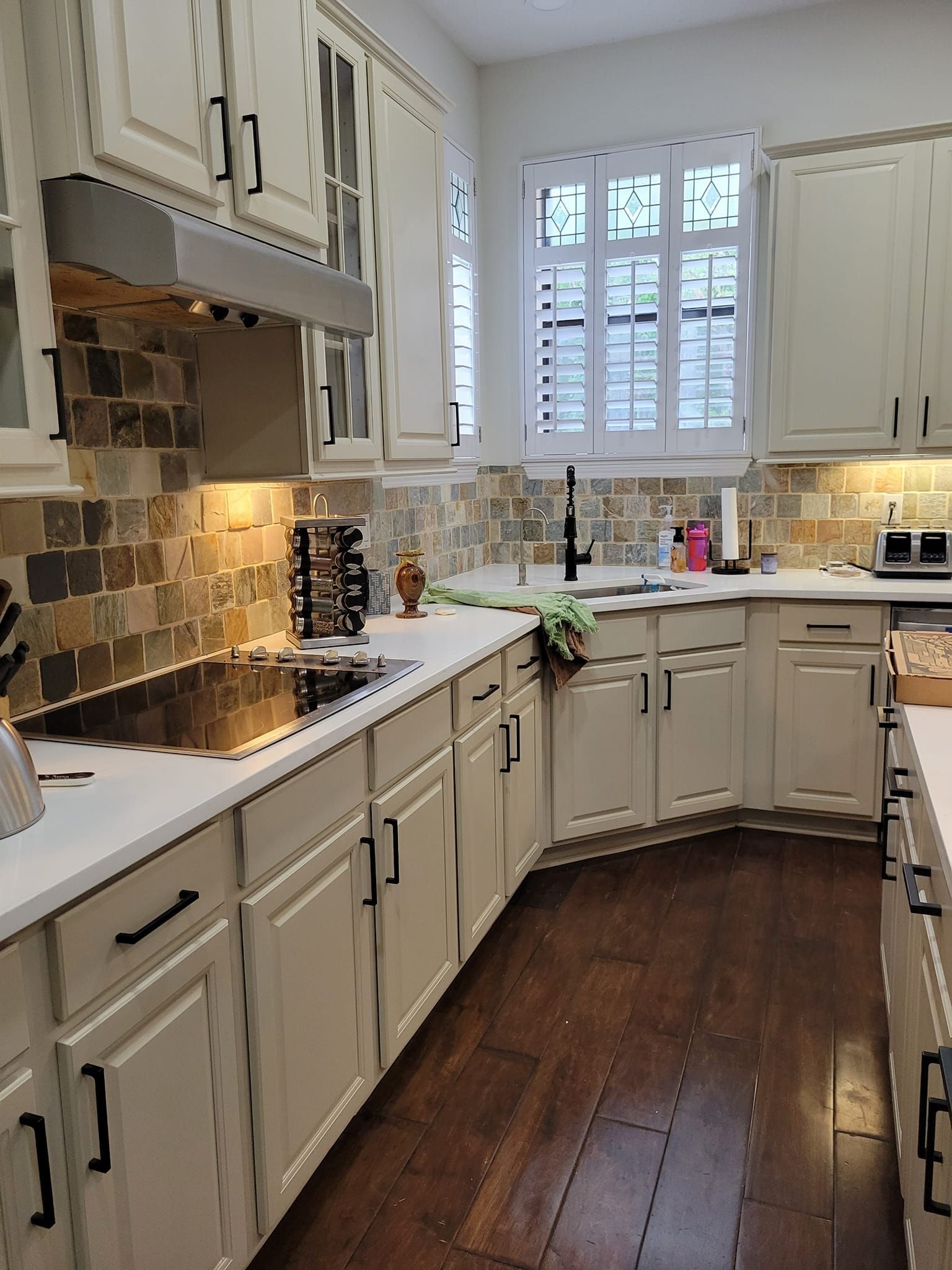 Cream-colored kitchen with dark wood floors and a tiled backsplash. Cabinets have black hardware and a window with white shutters is in the background.