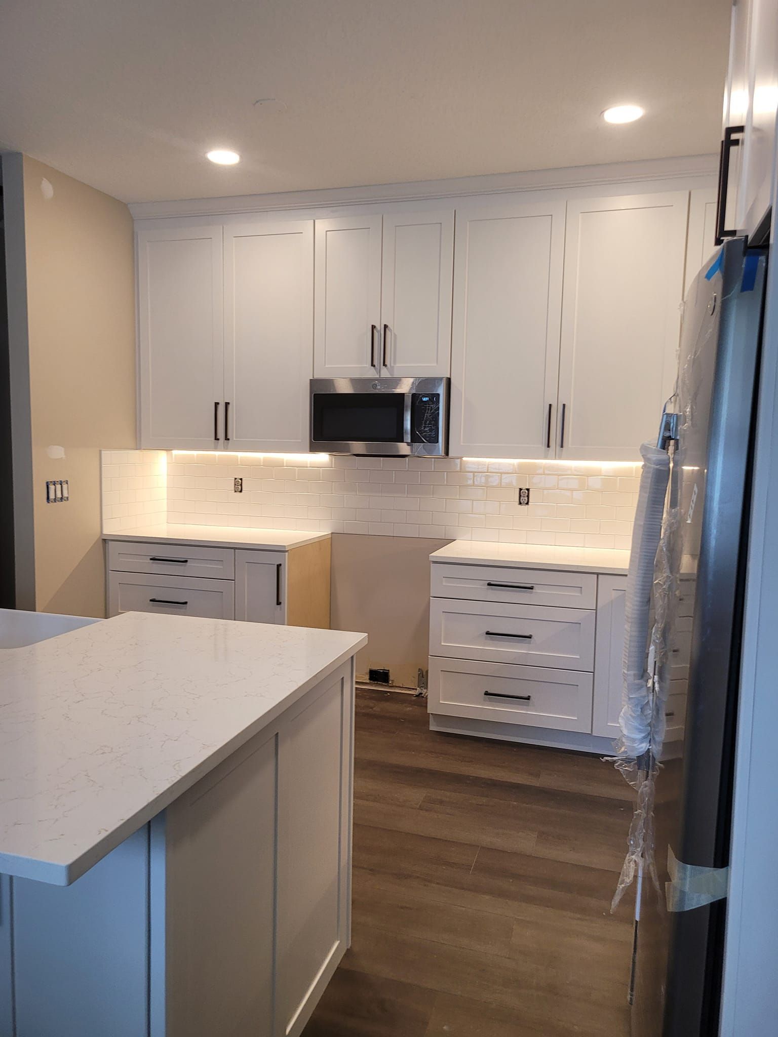 A newly renovated kitchen with white cabinets, a microwave, and a countertop. There's a kitchen island and a refrigerator on the right.