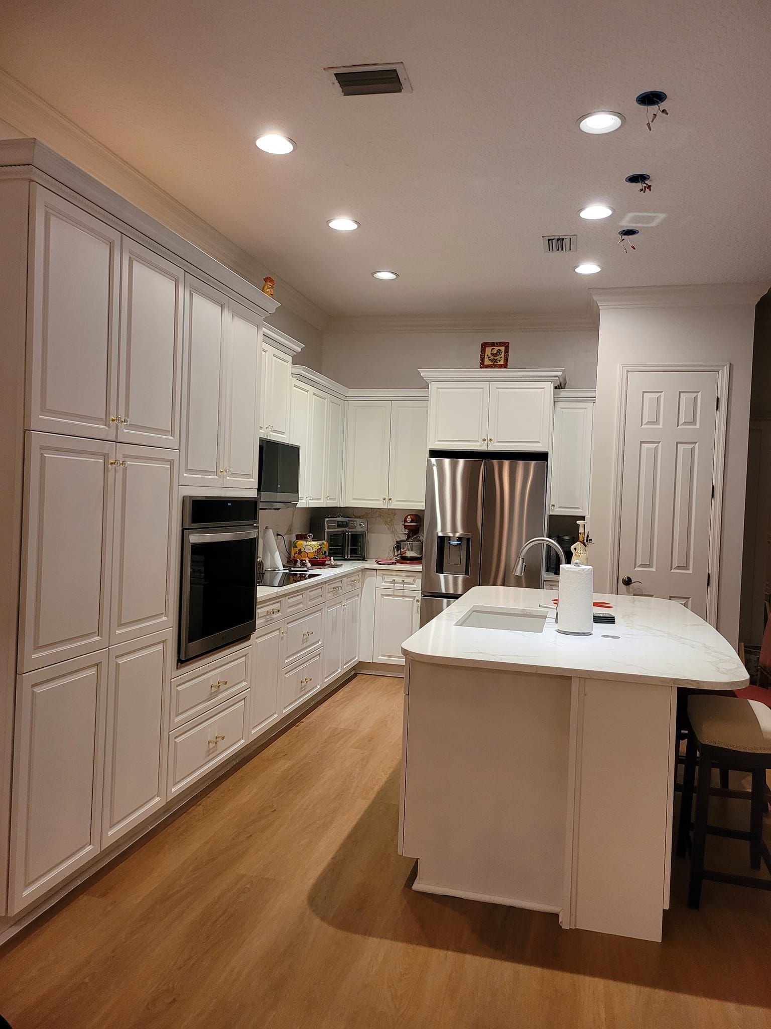 A well-lit kitchen with white cabinets, stainless steel appliances, and a wooden floor. There's an island with a white countertop.
