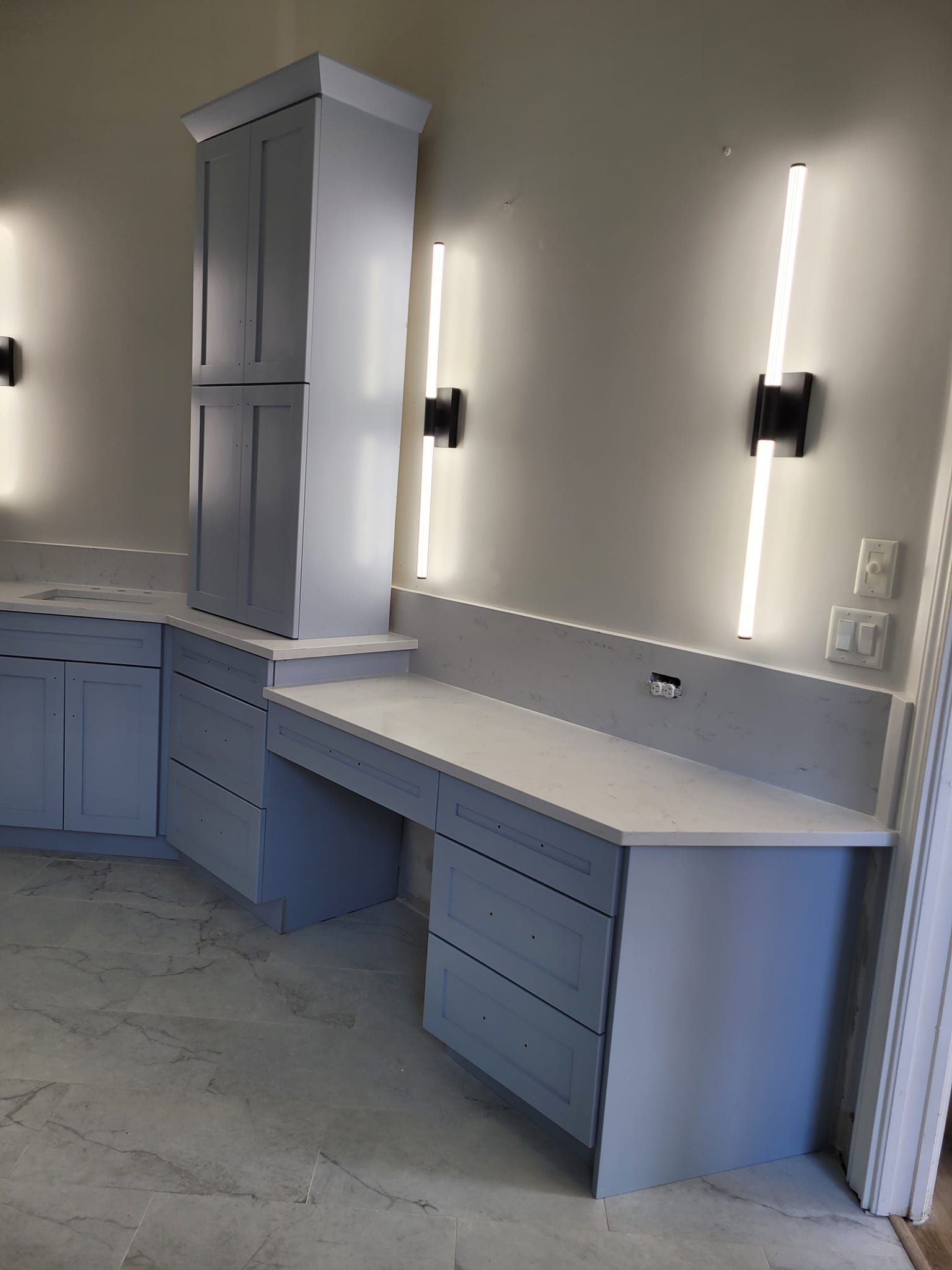 Built-in desk and cabinets painted light blue with white countertops, against a white wall with vertical lights and a black sconce.