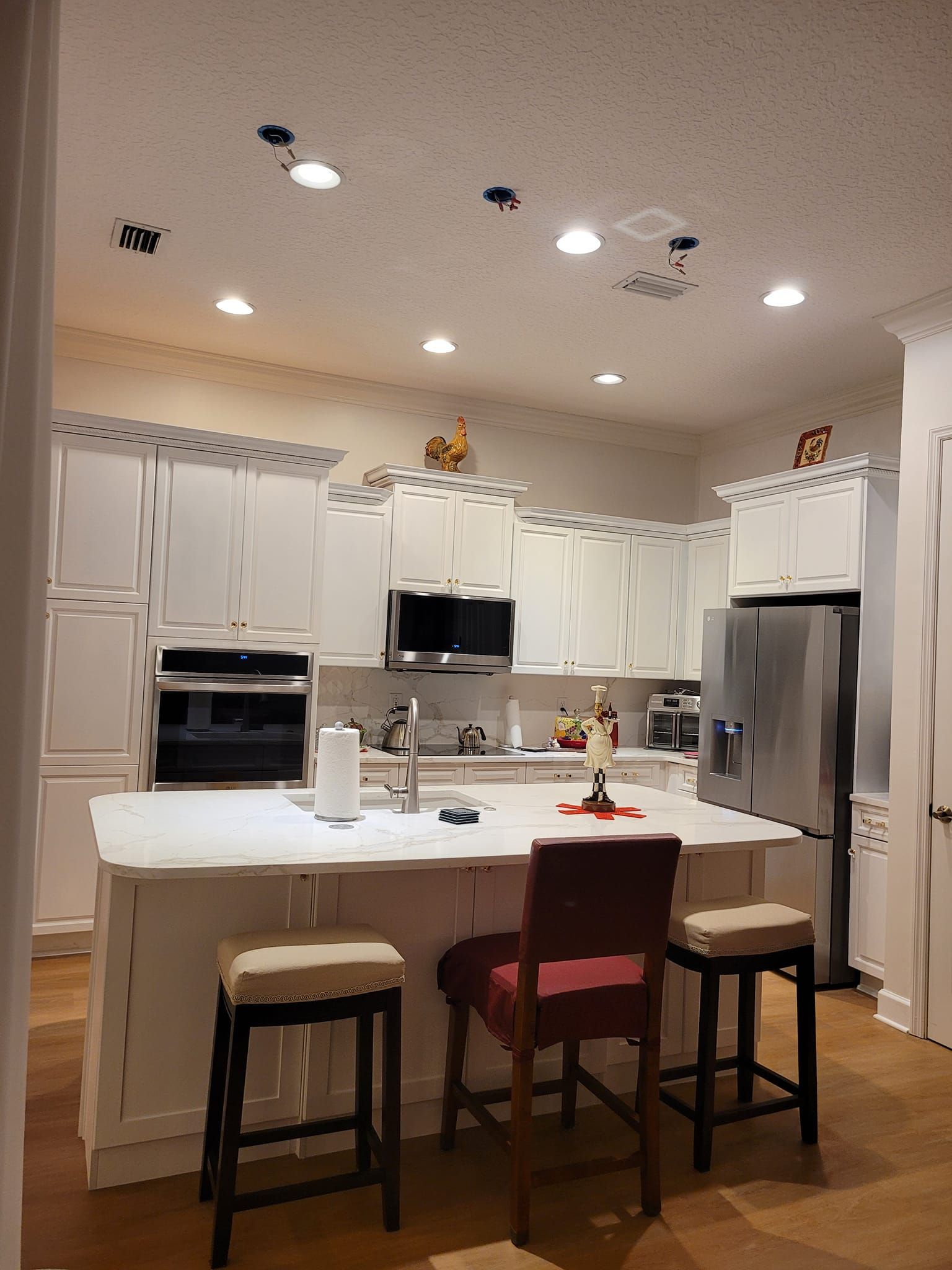 White kitchen with island, countertops, and stainless steel appliances. Overhead lighting and bar stools are present.