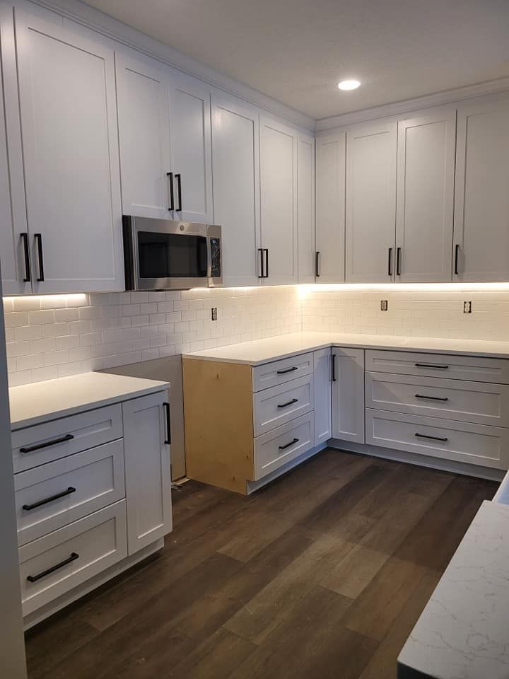 White kitchen cabinets with black hardware and a stainless steel microwave, white tiled backsplash, wood floors.