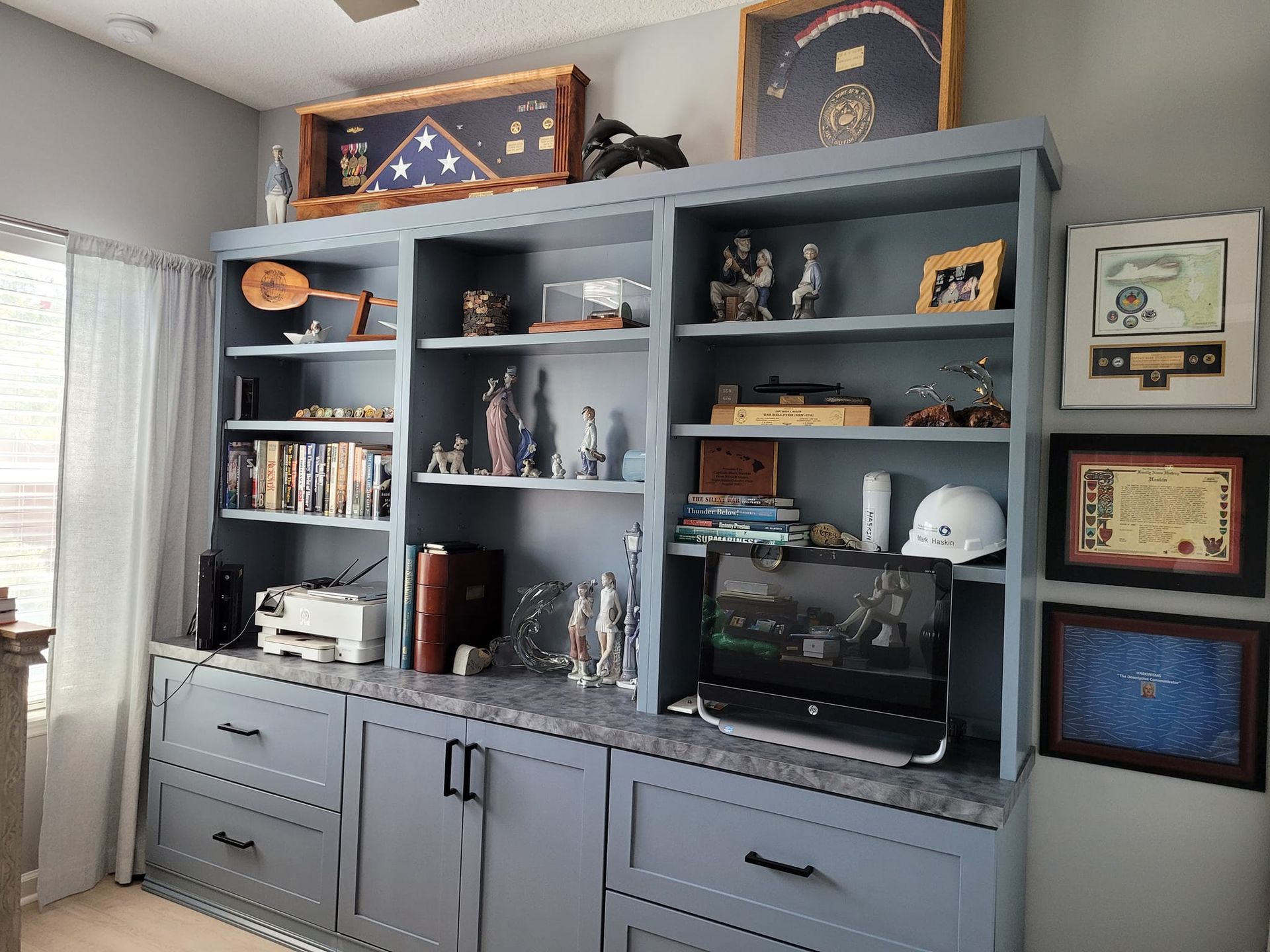 Blue built-in shelving unit with a desk area, holding books, decor, and a computer monitor. Several framed items adorn the wall.