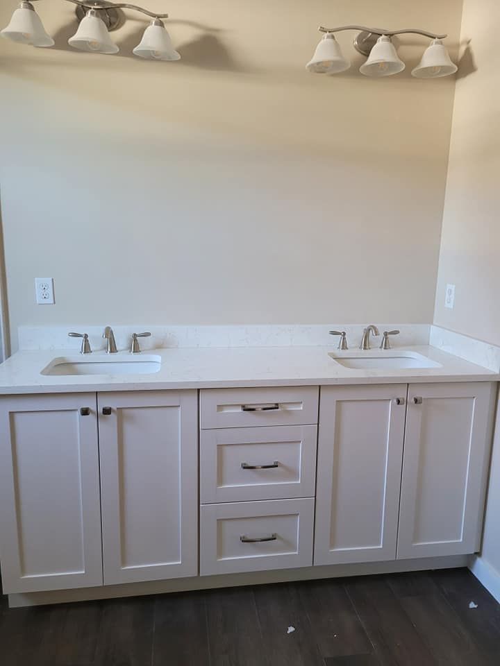 White bathroom vanity with two sinks, cabinets, and drawers. Above, are two light fixtures and a light tan wall.