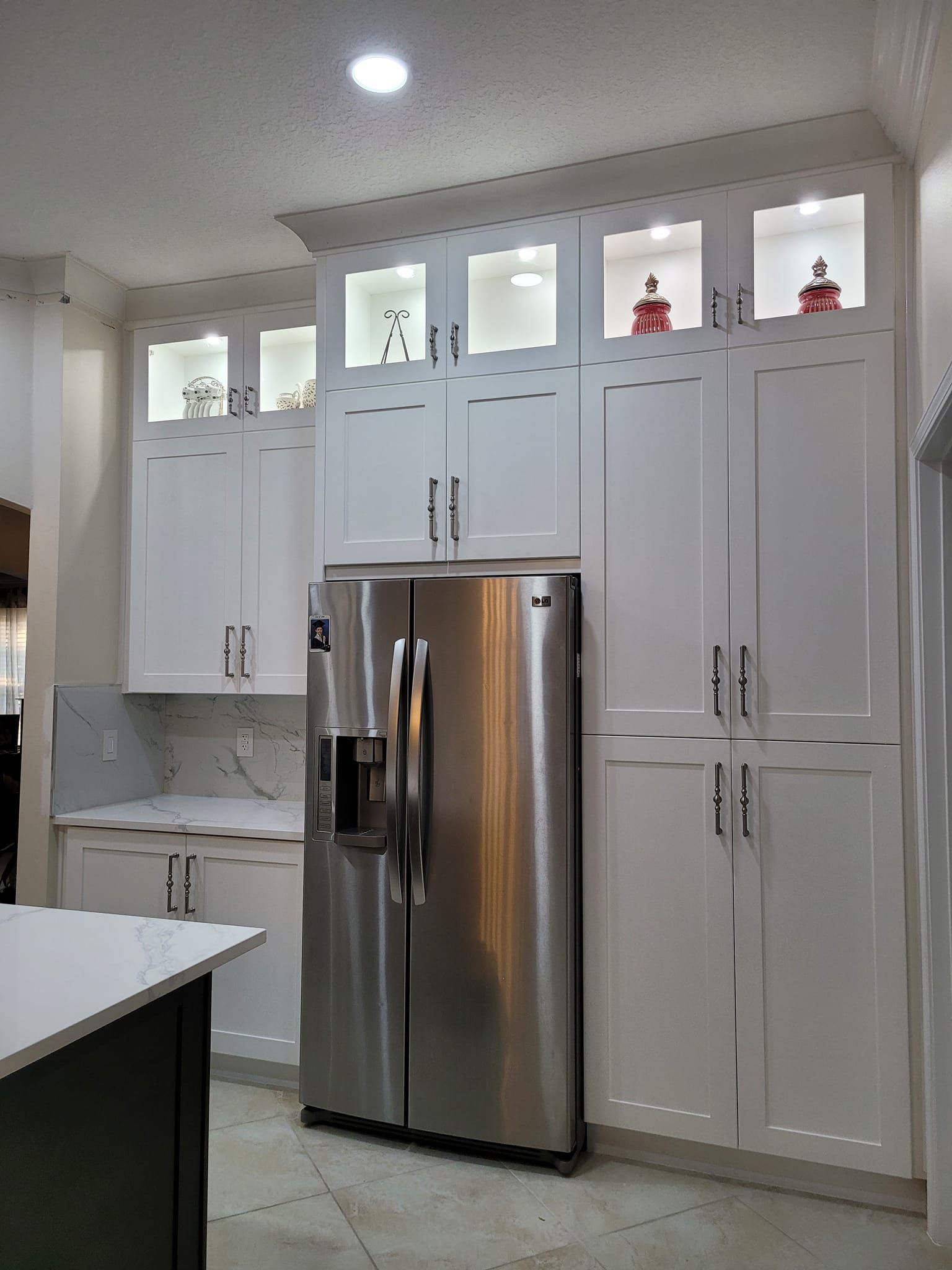 White kitchen cabinets surrounding a stainless steel refrigerator. Lighted display shelves sit atop the upper cabinets.