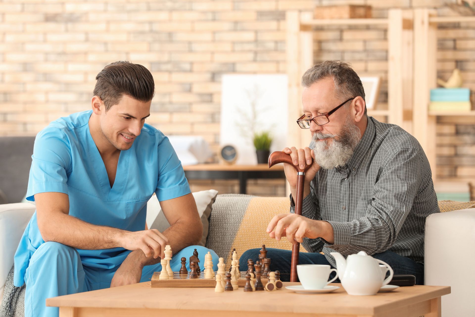 Man in blue scrubs plays chess with older man, who holds a cane and tea cup, at a table indoors.