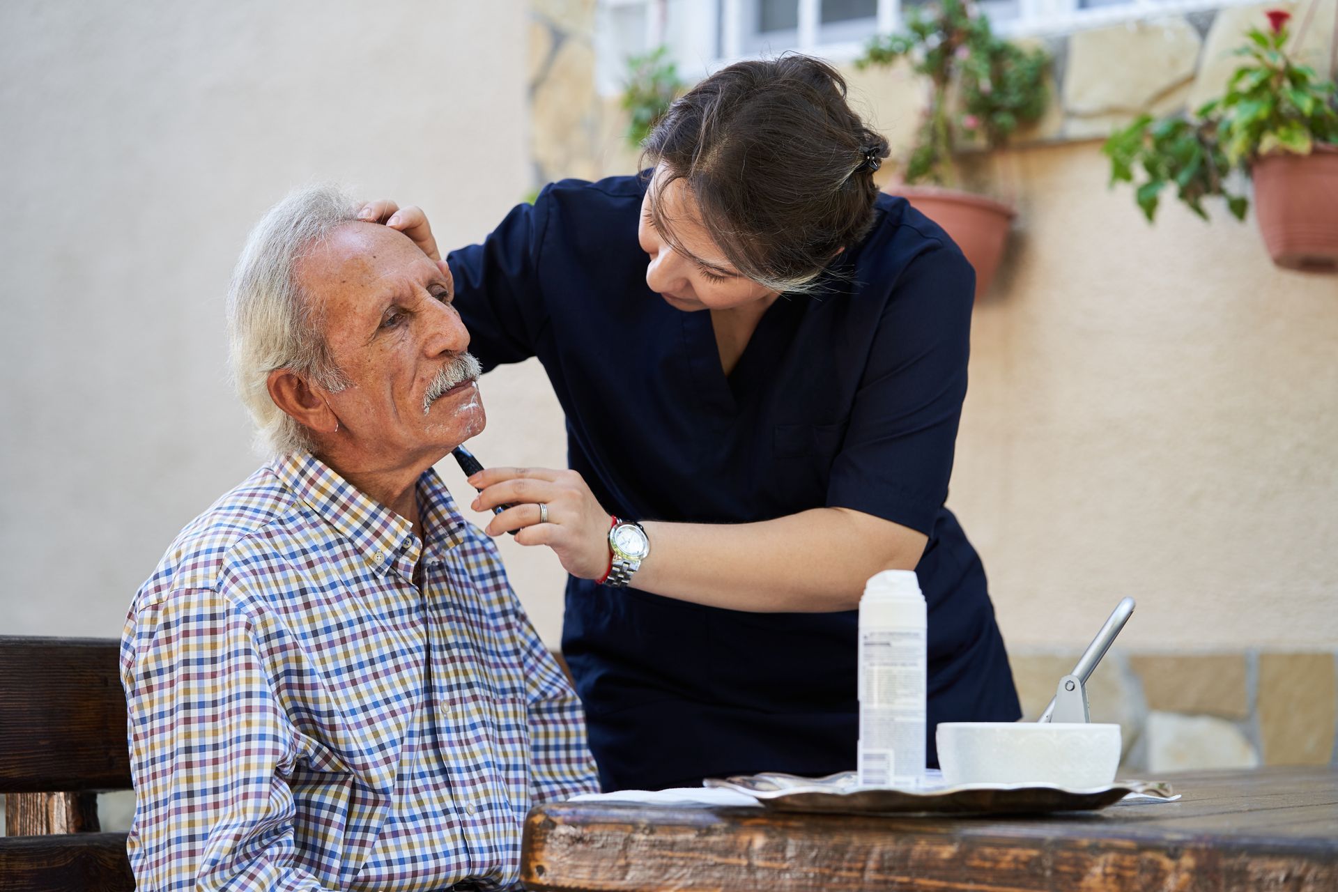 A person shaves the face of an older adult seated outdoors at a table.
