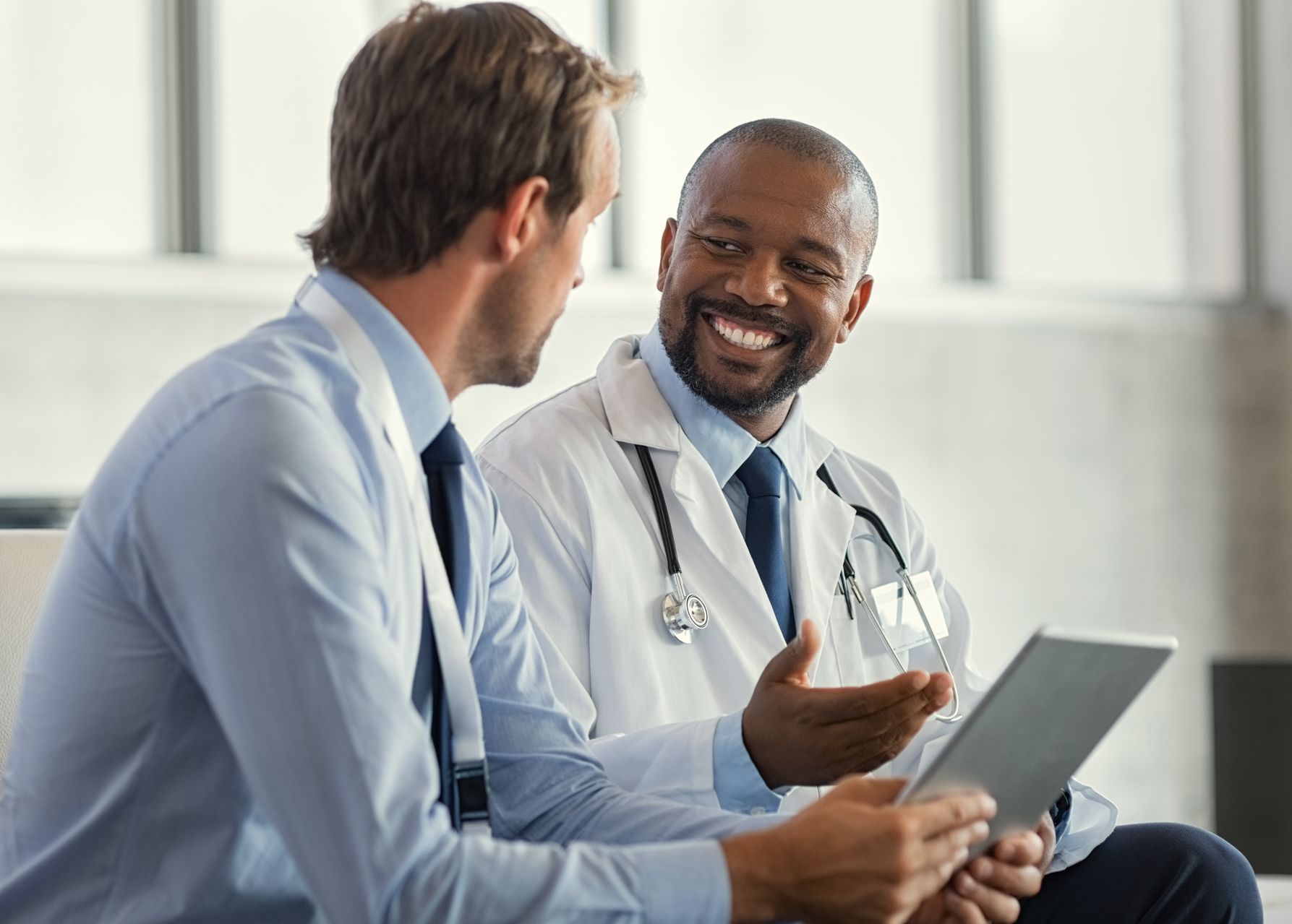 Doctor and patient looking at a tablet, smiling, indoors. The doctor is wearing a lab coat.