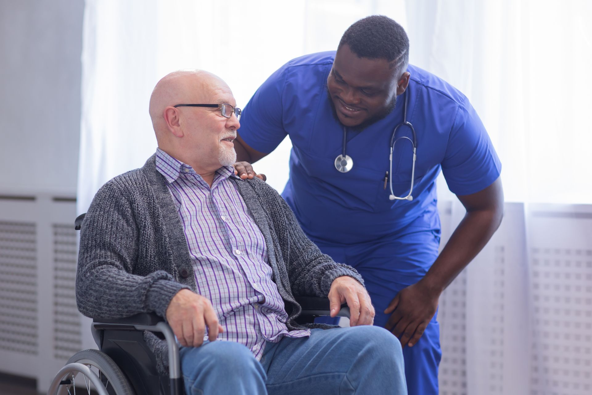 Man in wheelchair talking with a smiling healthcare worker in blue scrubs indoors.