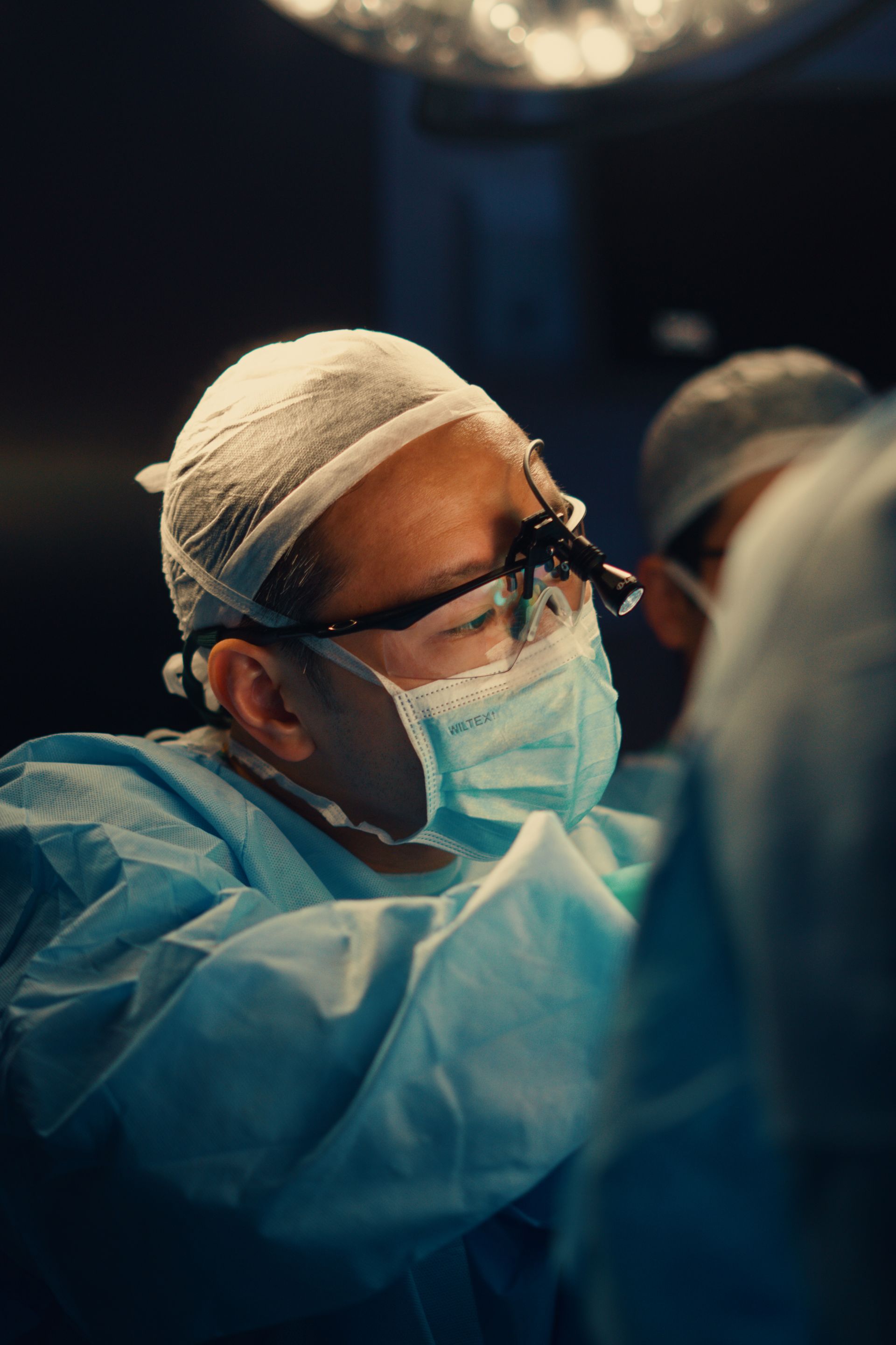 Cirurgião de uniforme azul, máscara e protetor facial realizando um procedimento em uma sala de cirurgia.