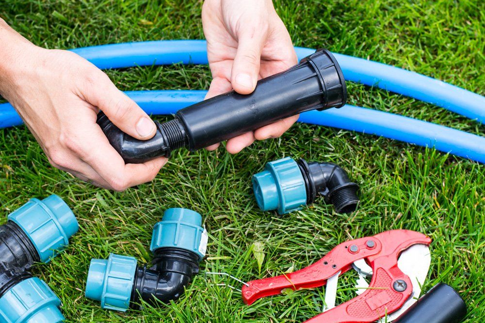 Person assembling black irrigation pipe fittings on grass, with blue hose and cutting tool.