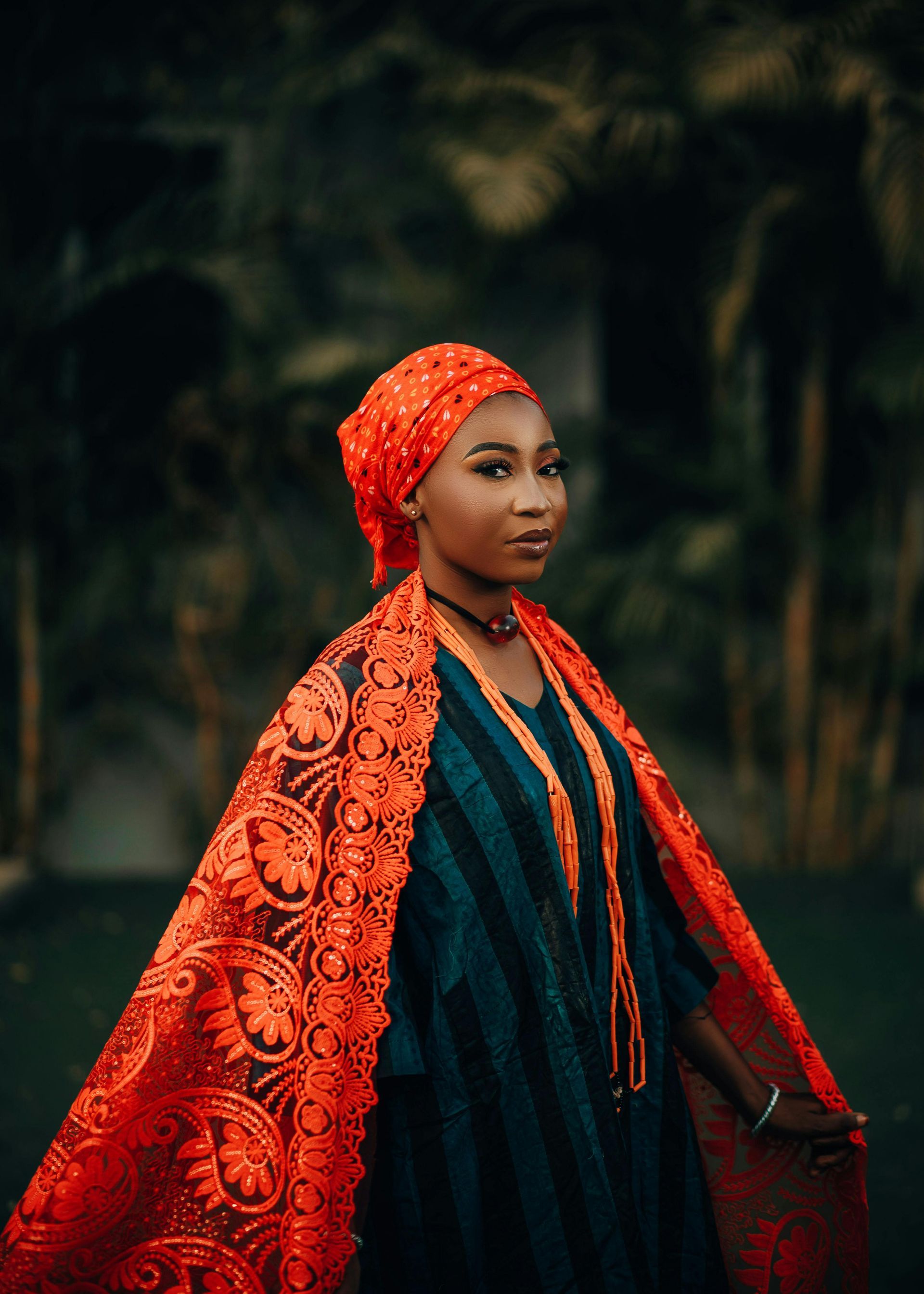 A woman wearing a turban and an orange cape is standing in front of trees.