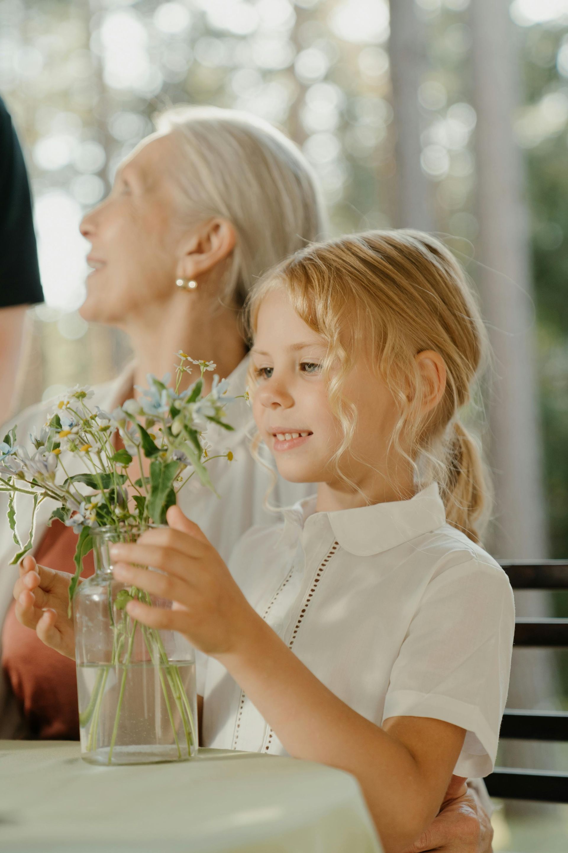 A little girl is sitting at a table holding a vase of flowers.