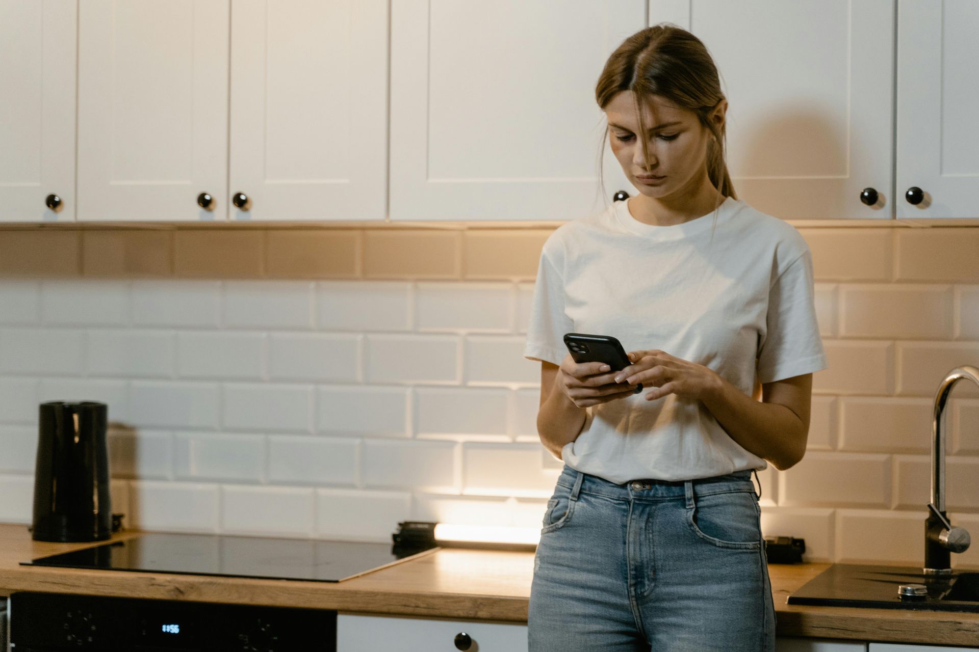 A woman is standing in a kitchen looking at her cell phone.