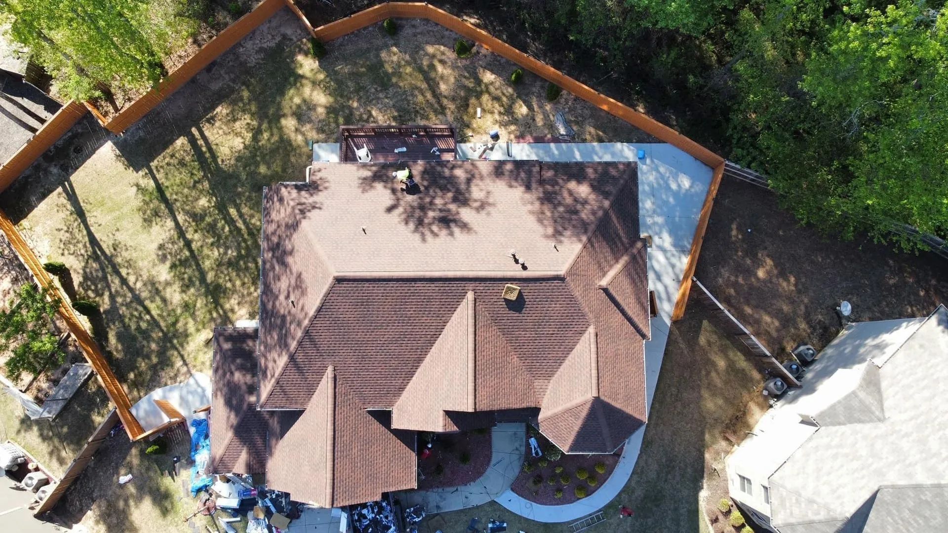 Overhead view of a house with a brown roof and a fenced-in yard, surrounded by trees and a paved driveway.
