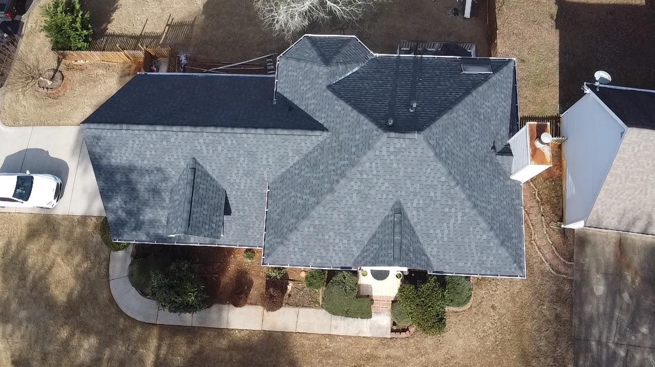 Overhead view of a house with gray roof, a driveway, and landscaping. A car is parked in the driveway.
