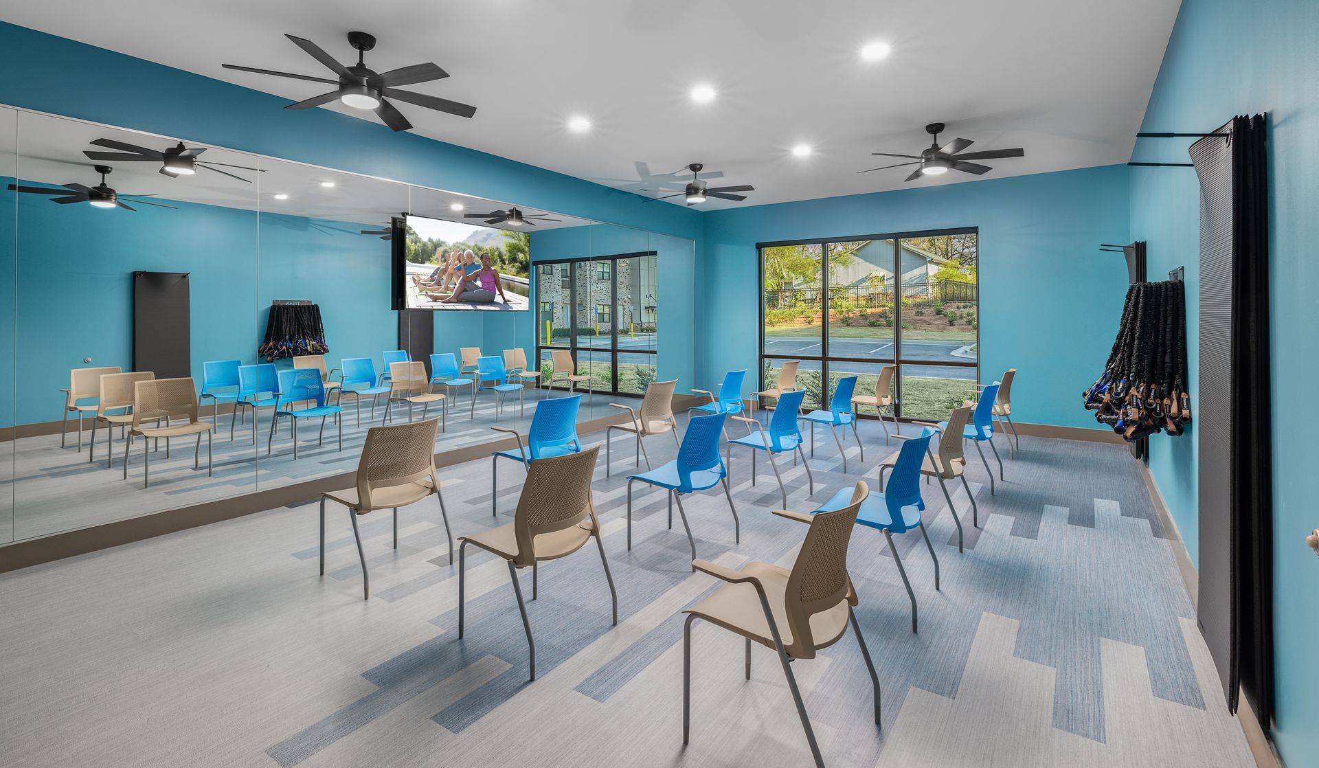 Bright meeting room with rows of blue chairs, tables, ceiling fans, and large windows.