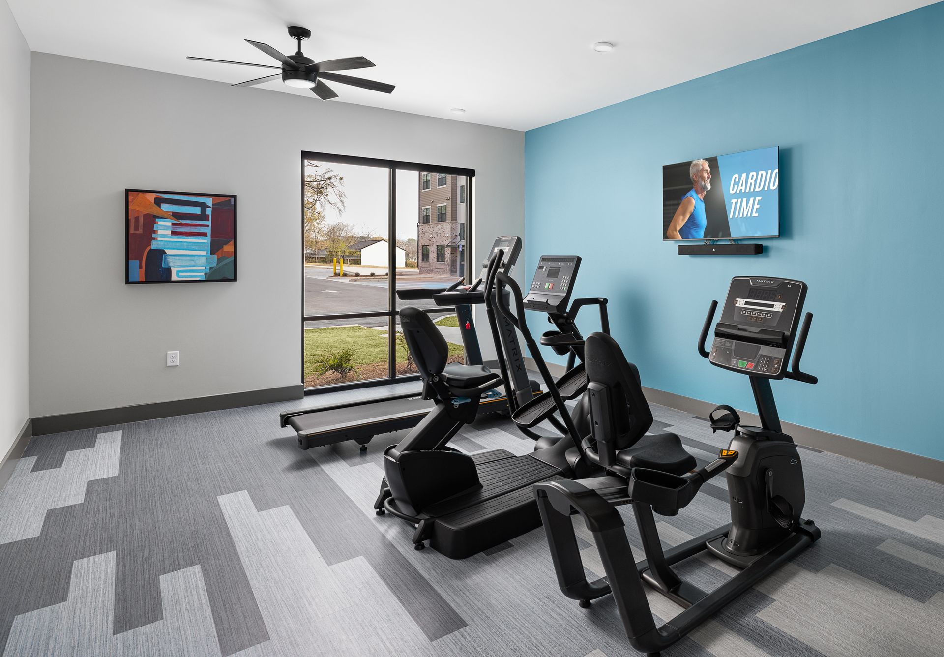 Bright gym room with cardio machines, gray carpet, blue and gray walls, and wall-mounted TVs