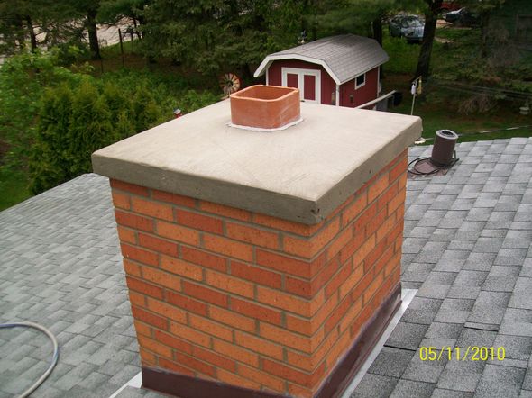 A person in work clothes inspects a red brick chimney on a shingled roof under a clear blue sky.