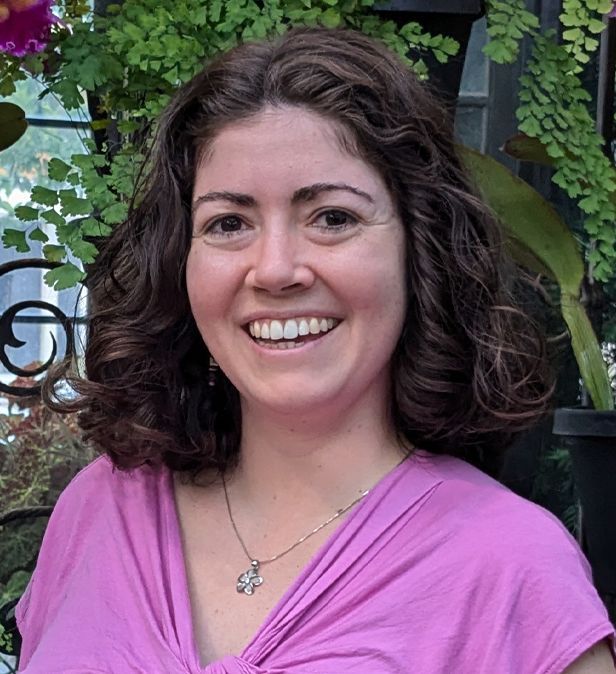 Smiling woman in a pink top standing outdoors with leafy greenery behind her