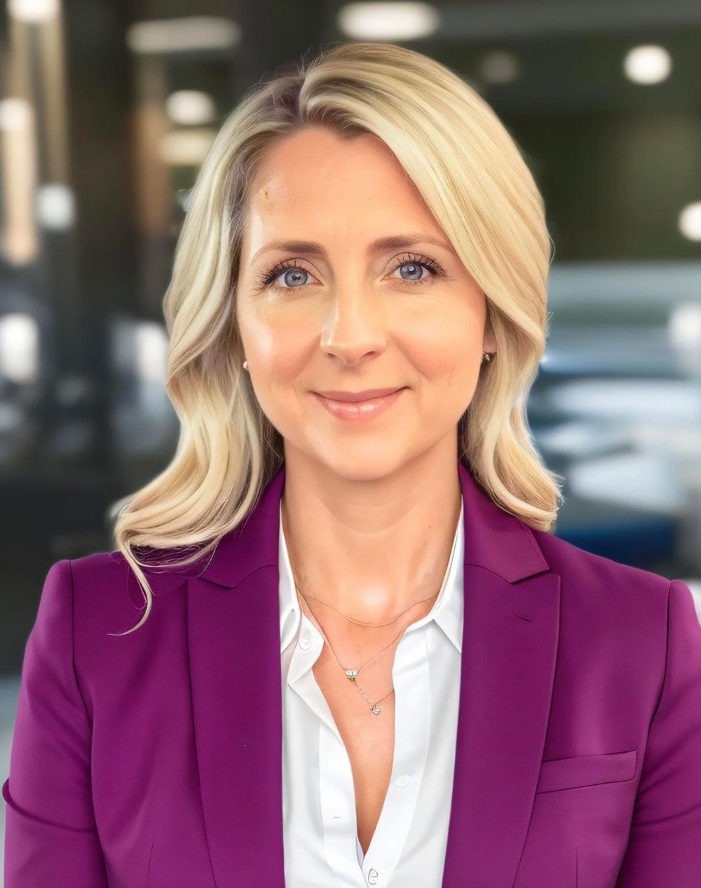 Woman with blonde hair in a purple blazer, smiling, wearing a white shirt, and a necklace. Blurred office setting.