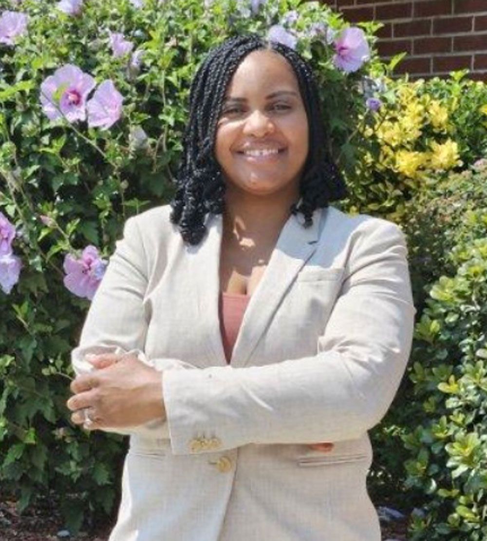 A woman in a suit is standing in front of a bush with purple flowers.