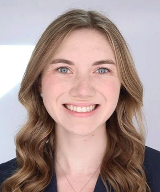Woman with light brown hair smiles, wearing a navy blazer, in front of a white background.