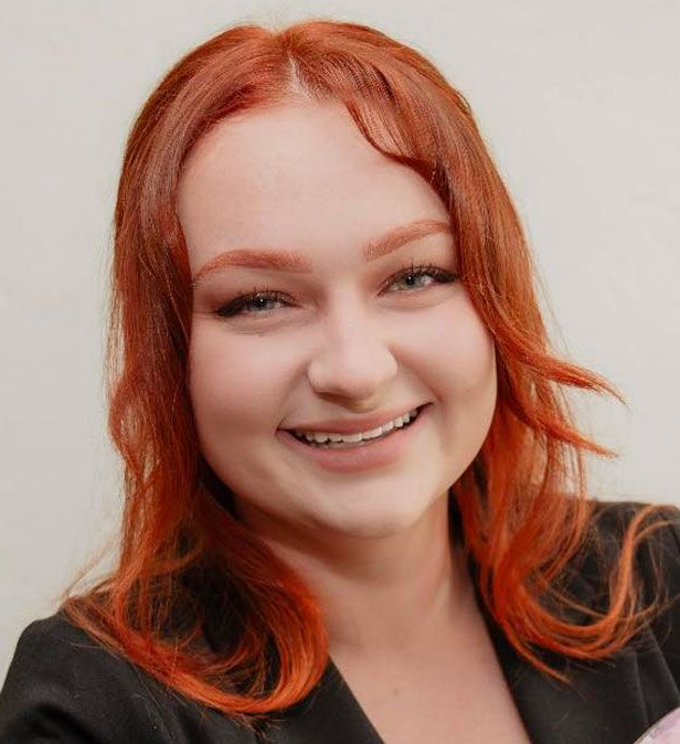 Woman with red hair smiling, holding paint palette and brushes, wearing a black blazer.