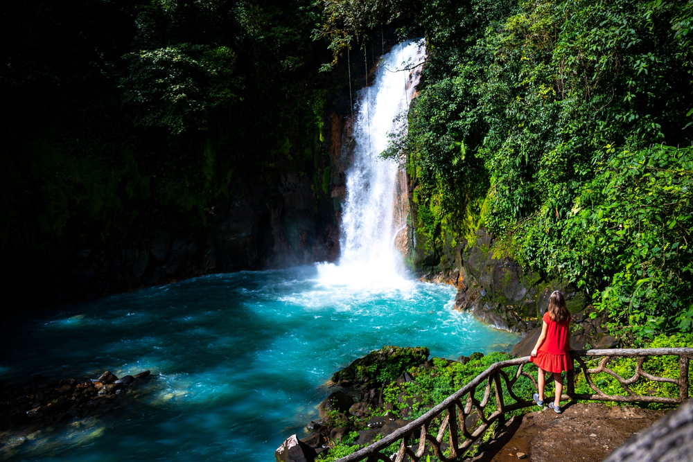 Woman in red dress overlooking a turquoise waterfall in lush green rainforest.
