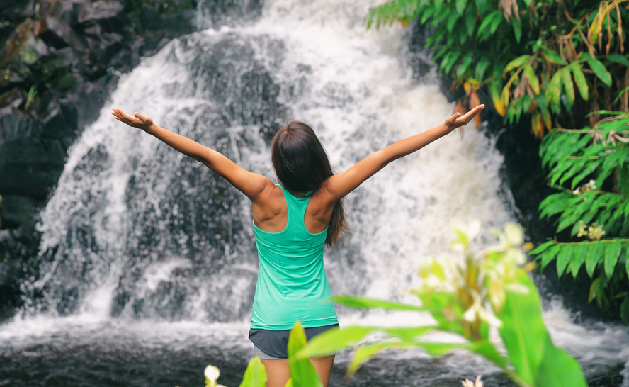 A woman is standing in front of a waterfall with her arms outstretched.