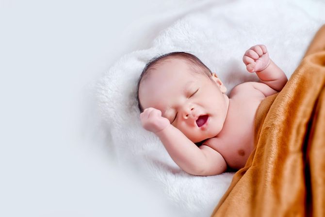 A newborn baby is sleeping on a white blanket with its mouth open.