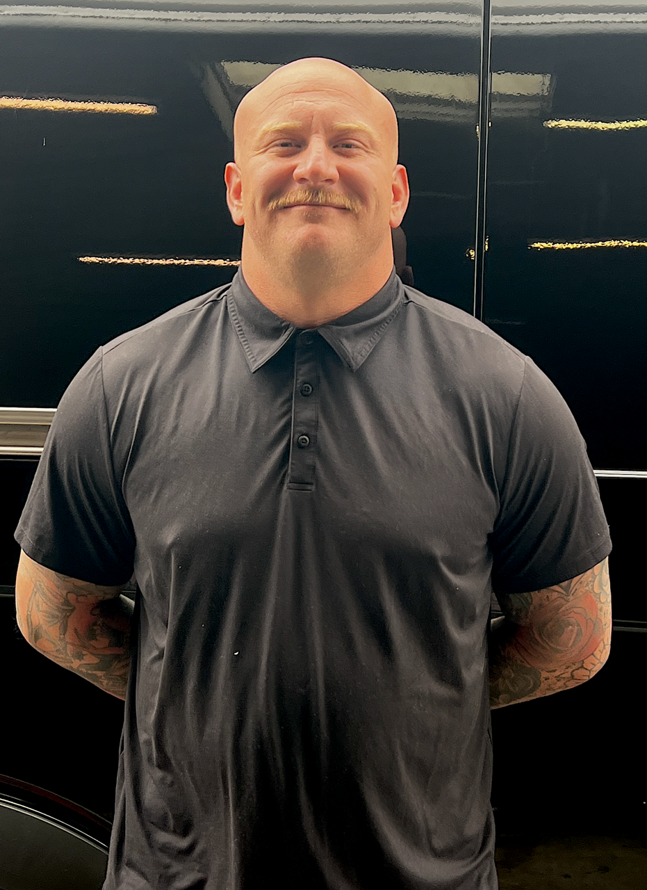 Smiling bald man in a dark polo shirt standing with hands behind his back against a black wall