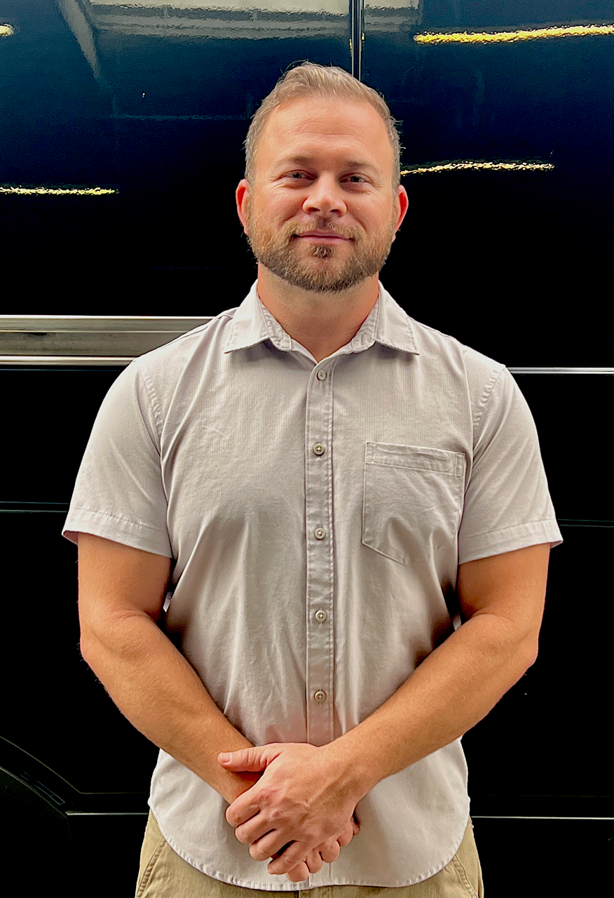 Man standing indoors against a dark wall, wearing a light gray short-sleeve button-up shirt.