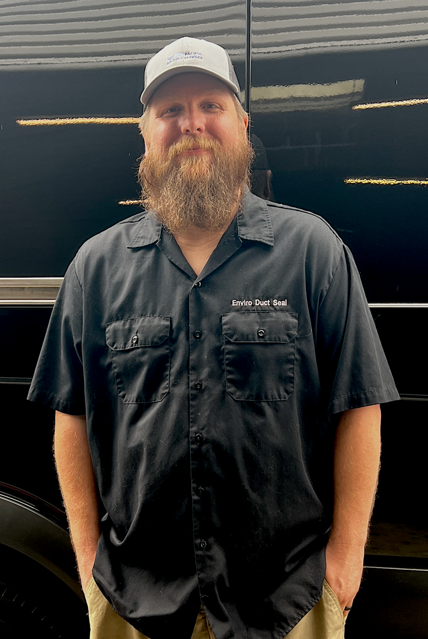 Man in a gray cap and dark shirt standing in front of a black vehicle, smiling.