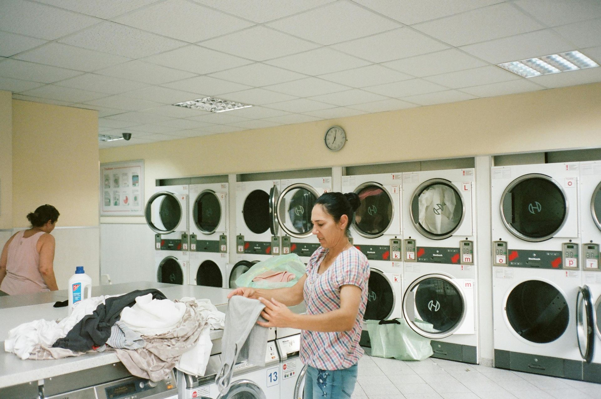Laundry room with rows of dryers; two people folding clothes near a pile of laundry.