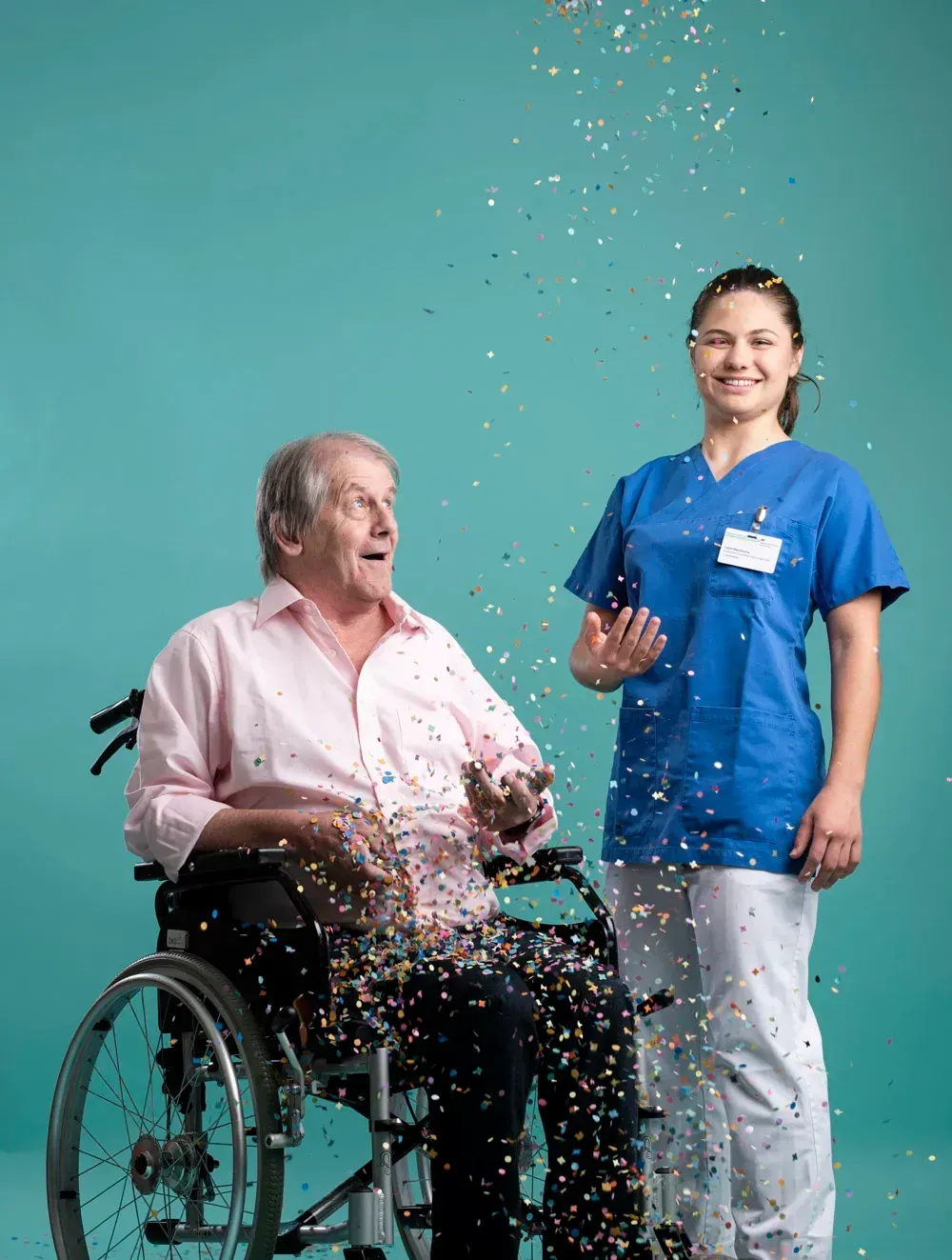 aelterer Herr im Rollstuhl mit Pflegerin, werfen Konfetti und lachen, dezentes Make-Up