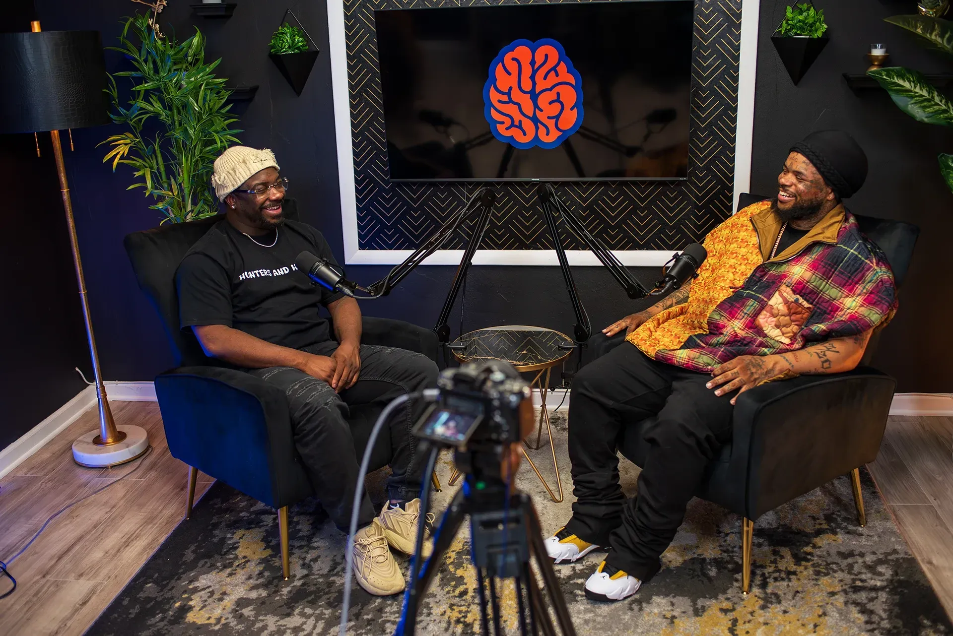 Three men in a gallery. One speaks into a mic while seated, two stand by paintings. Graffiti art background.